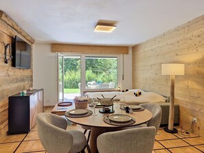 A cozy room with a dining table set for four, a bed with pillows, a TV mounted on the wall, and a window with a view of greenery. - Les Ambassadeurs B 3 by Interhome (Crans-Montana)