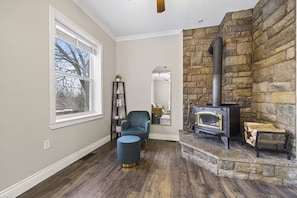 Une pièce avec une cheminée en pierre, une fenêtre donnant sur les arbres, un fauteuil bleu et un plancher en bois.