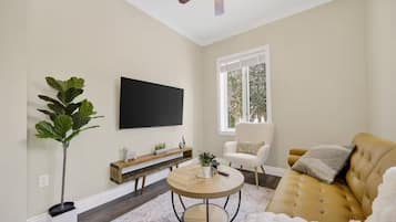 Un salon comprenant un téléviseur à écran plat, une table basse ronde, un canapé, un fauteuil, une plante et une fenêtre donnant sur des arbres.