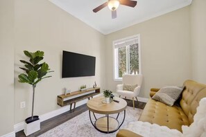 A living room with a flat-screen TV, a round coffee table, a sofa, a chair, a plant, and a window with a view of trees.