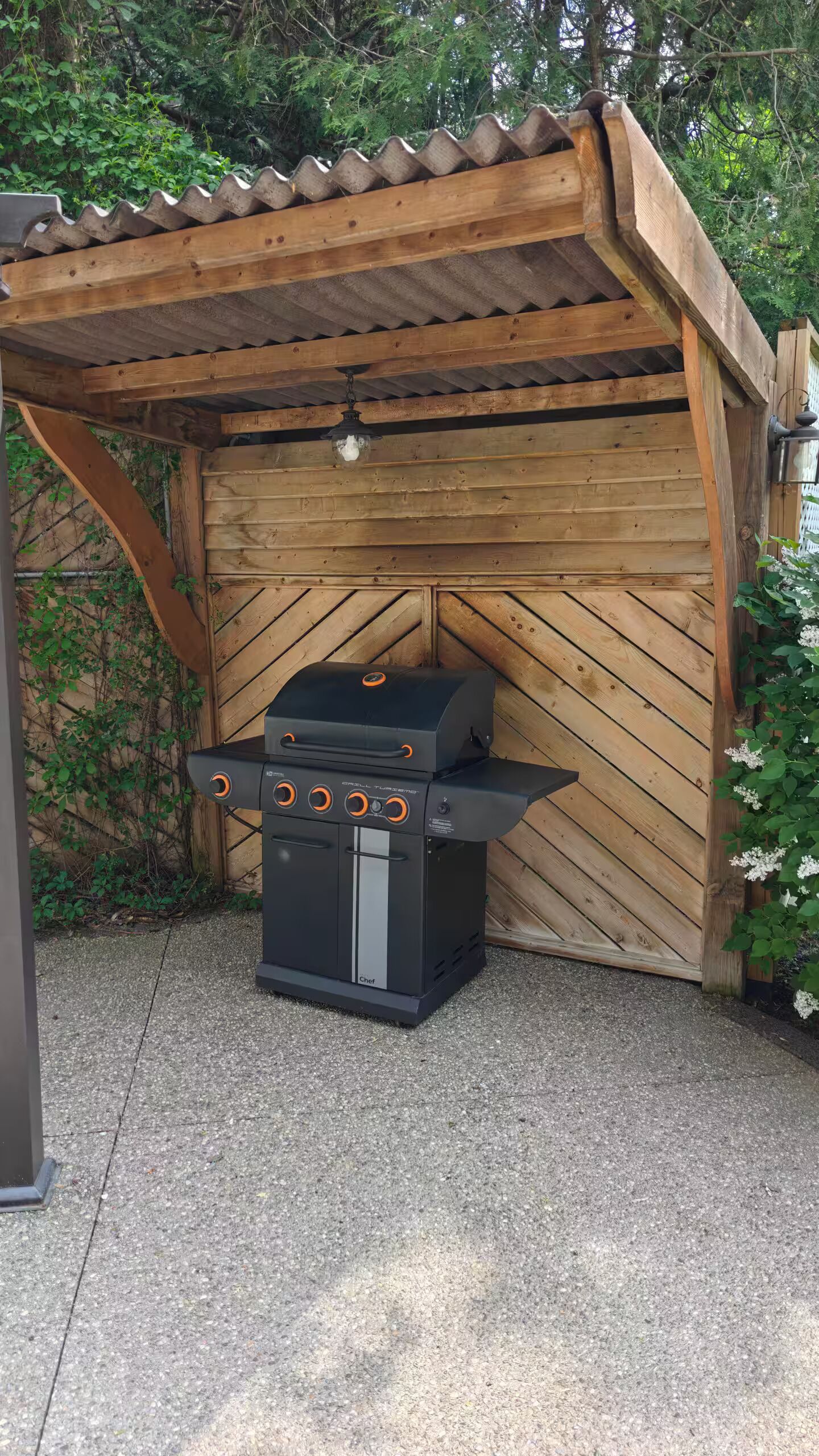 A barbecue grill under a wooden pergola with a sloped roof, surrounded by greenery.