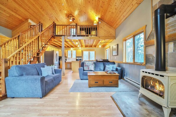 A spacious living area with a wooden staircase, a fireplace, and a kitchen in the background.