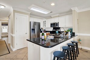 A modern kitchen with white cabinetry, a stainless steel refrigerator, a microwave, and a black countertop. There are three black bar stools and a small island with a bottle of champagne and glasses. - Colorful Oceanfront Escape on Tybee's South End | Heated Pool Access (Tybee Island)