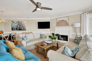 A living room with a ceiling fan, a fireplace, a TV mounted on the wall, and a coffee table with decorative items. - Colorful Oceanfront Escape on Tybee's South End | Heated Pool Access (Tybee Island)