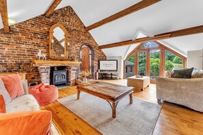 A spacious living room with exposed wooden beams, a brick fireplace, and a large window offering a view of the outdoors. - Luxury Stay Sleeps 14 Near M 6 (Newcastle Under Lyme)