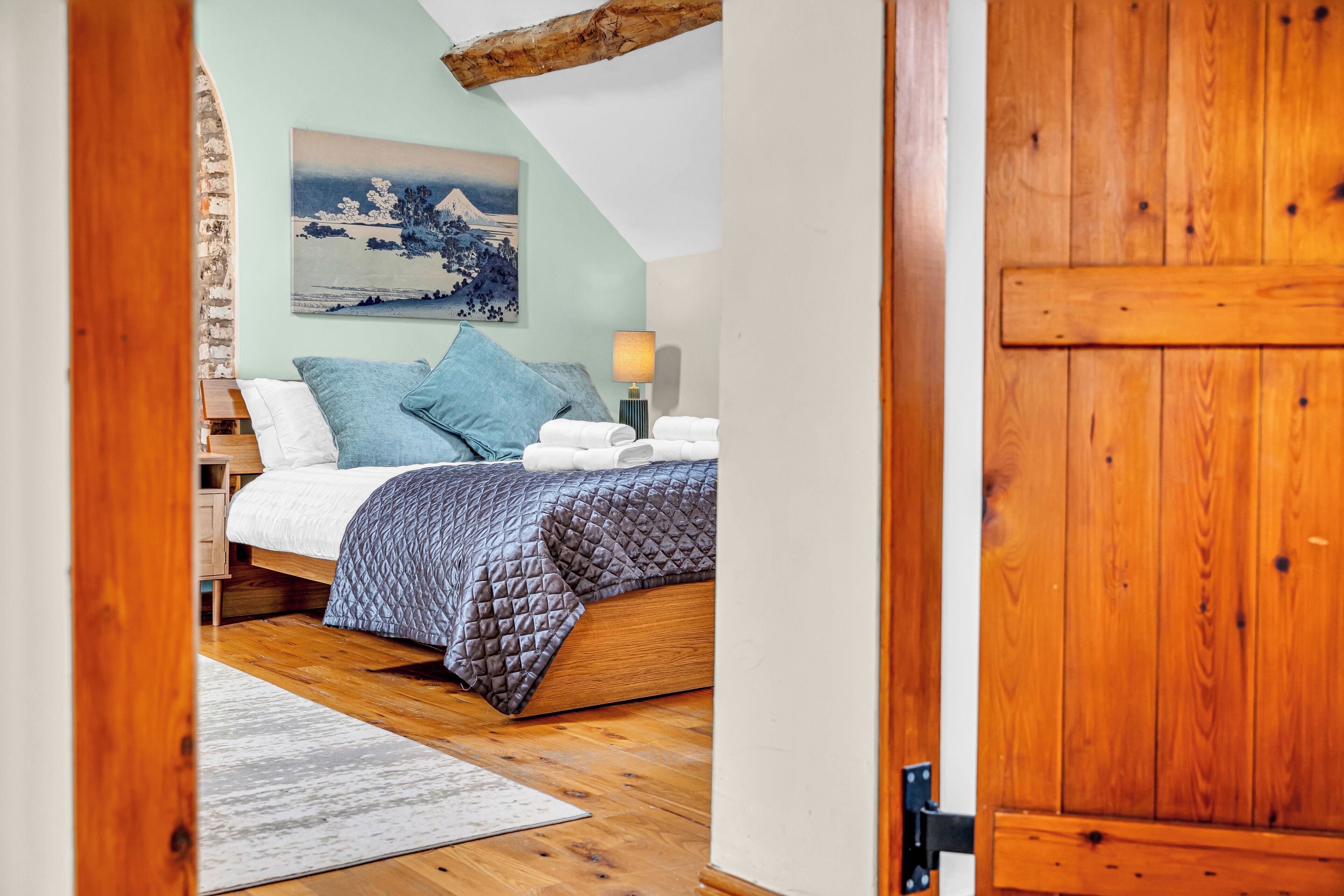 A bedroom with a bed, pillows, a bedside table, a lamp, and a painting on the wall.