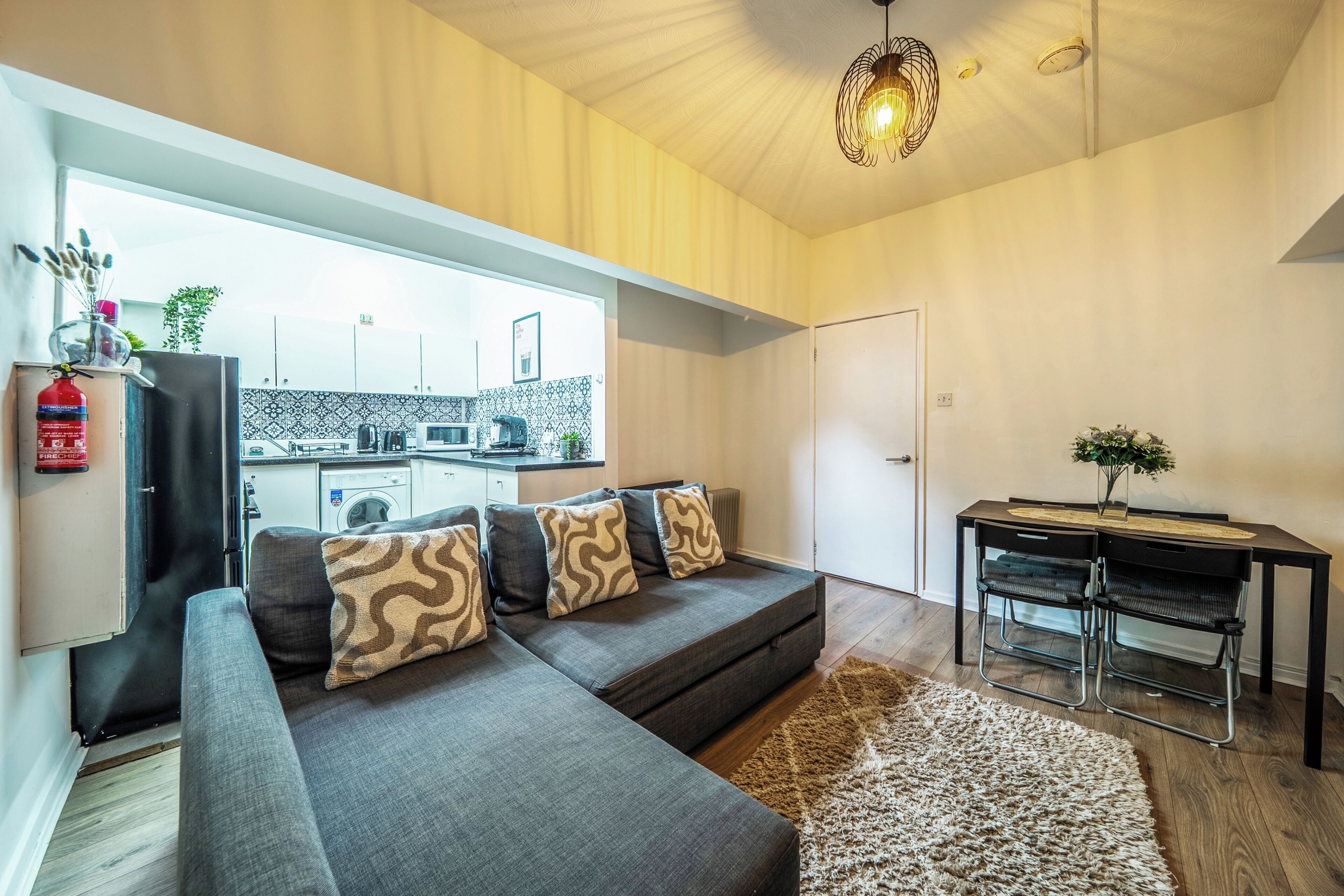 A modern living room with a grey sofa, a dining table with chairs, and a kitchen in the background.