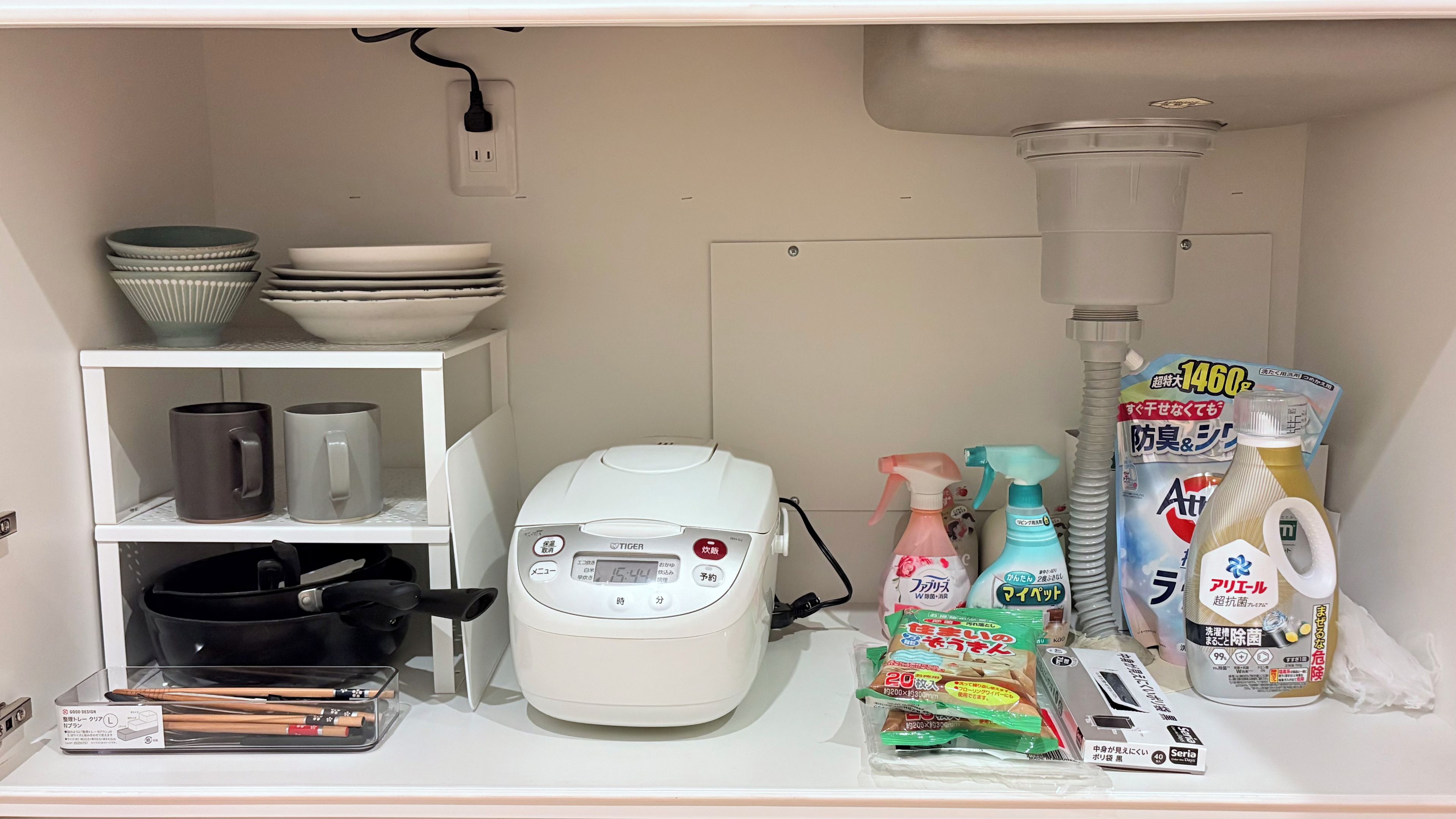 A kitchen area with a rice cooker, bowls, mugs, cleaning supplies, and a trash can.