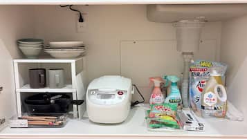 A kitchen area with a rice cooker, bowls, mugs, cleaning supplies, and a trash can.