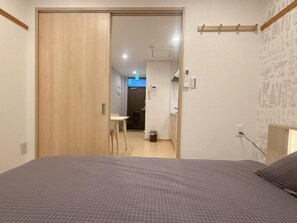 A bedroom with a bed, a wooden wardrobe, a small table, and a wall-mounted shelf. - Aoba Residence Designer Apartments 302 (大阪市)