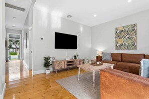 Un salón moderno con un televisor de pantalla plana, un sofá marrón, una mesa de centro de madera y una obra de arte con motivos de hojas tropicales en la pared.