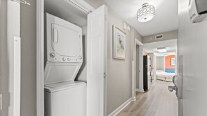 A laundry room with a front-loading washer and dryer, a hallway with a door leading to a bedroom, and a ceiling light fixture. - Oceanfront Penthouse, Pool, Lazy River & Waterpark (North Myrtle Beach)