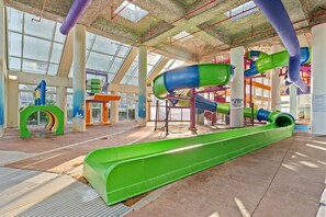 Een overdekt waterpark met een groene glijbaan, speelconstructies en een glazen dak.