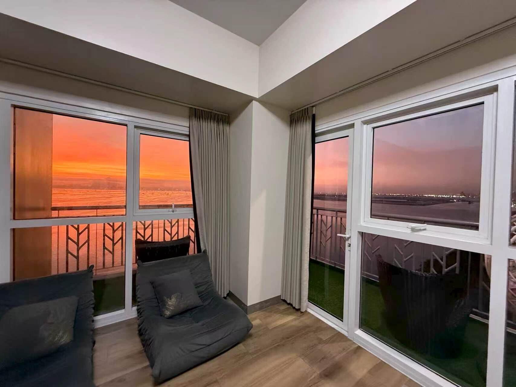 A balcony with a view of a sunset over the water, a cushioned seating area, and a glass door leading to an outdoor space.