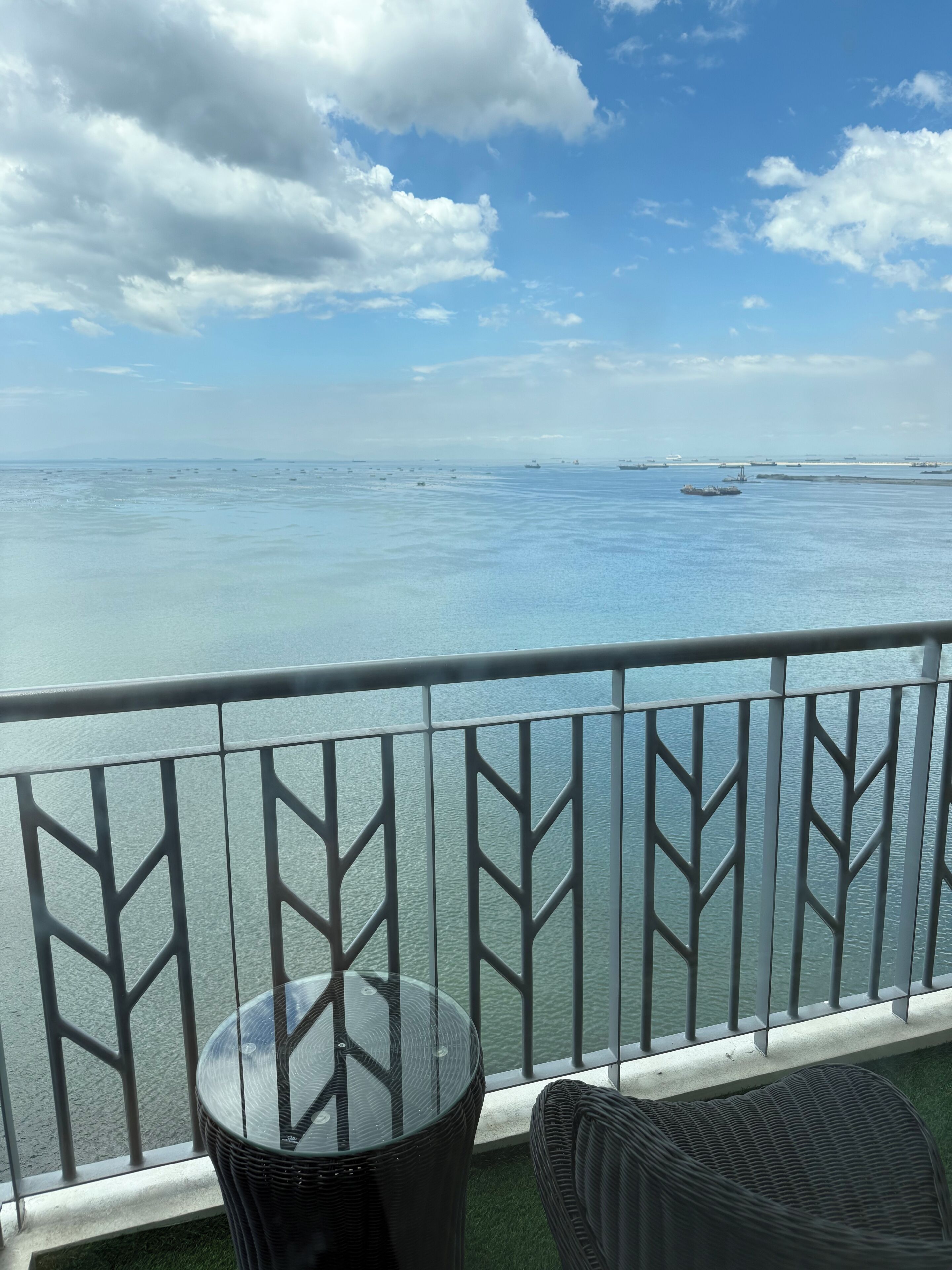 Ein Balkon mit Glastrennwand und Blick auf ein Gewässer, ein runder Tisch und ein Korbstuhl.