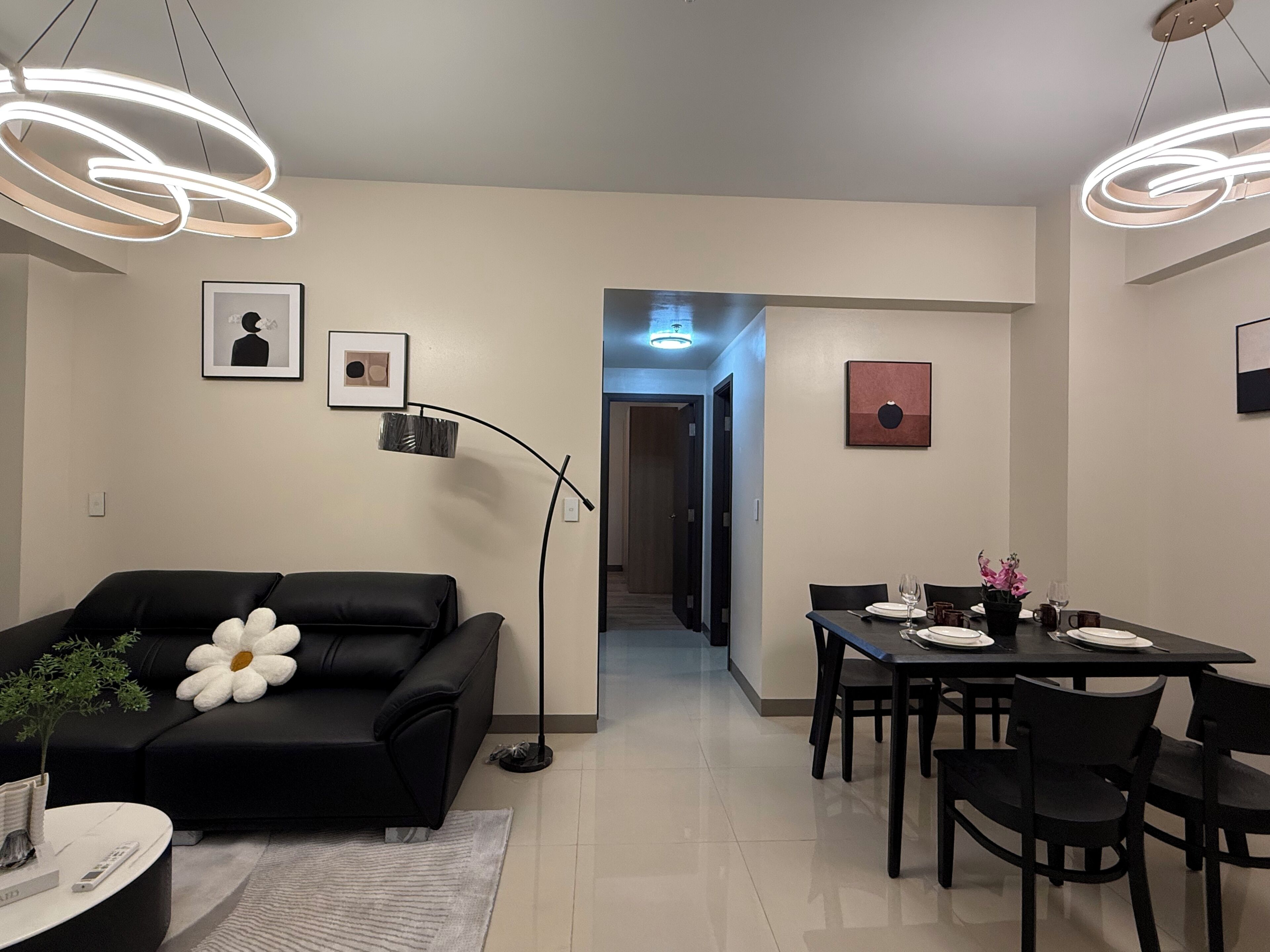 A modern living room with a black sofa, a dining table set for two, and a hallway leading to another room.