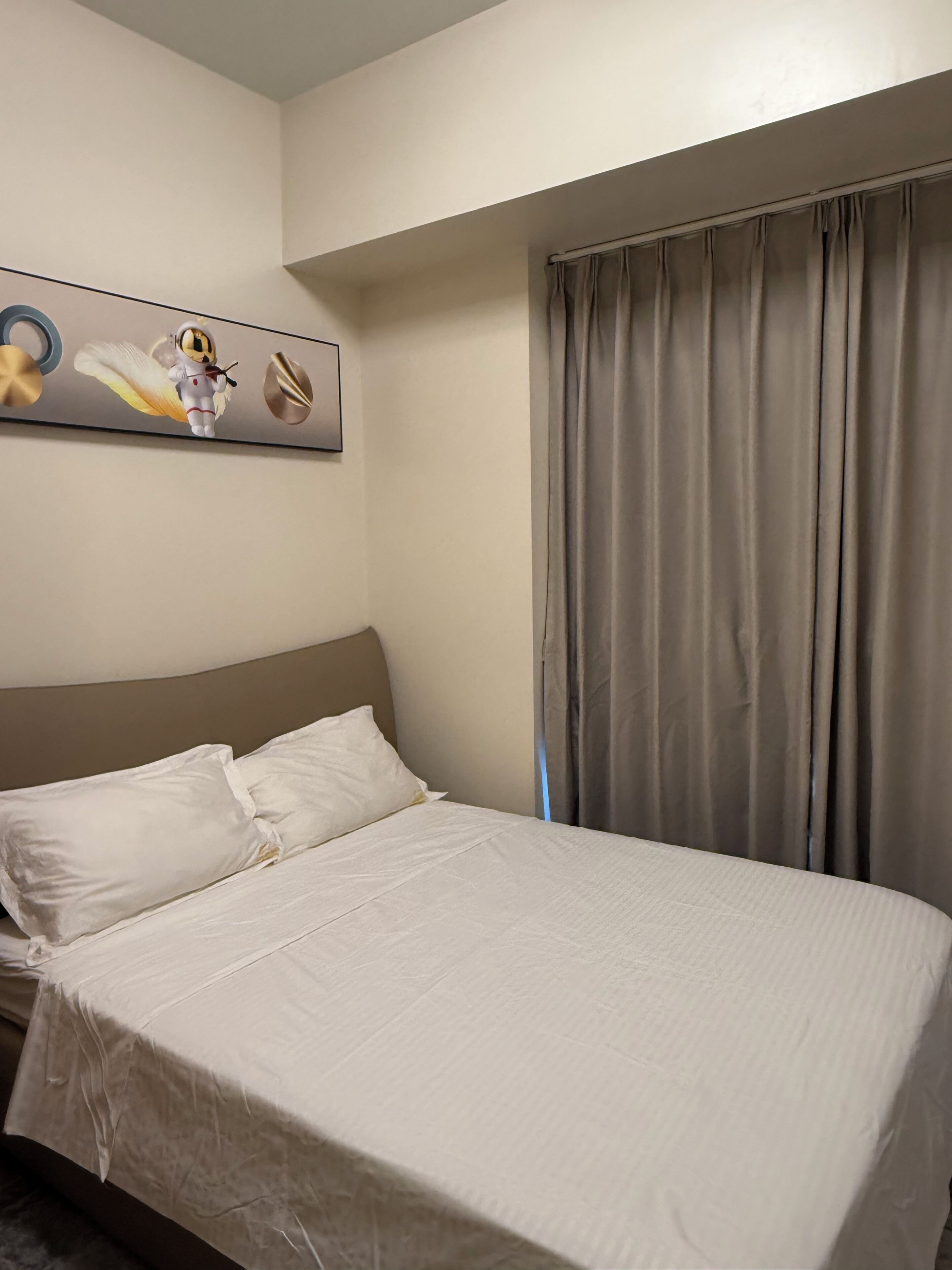 A hotel room with a bed, pillows, a headboard, a window with curtains, and a wall-mounted artwork.