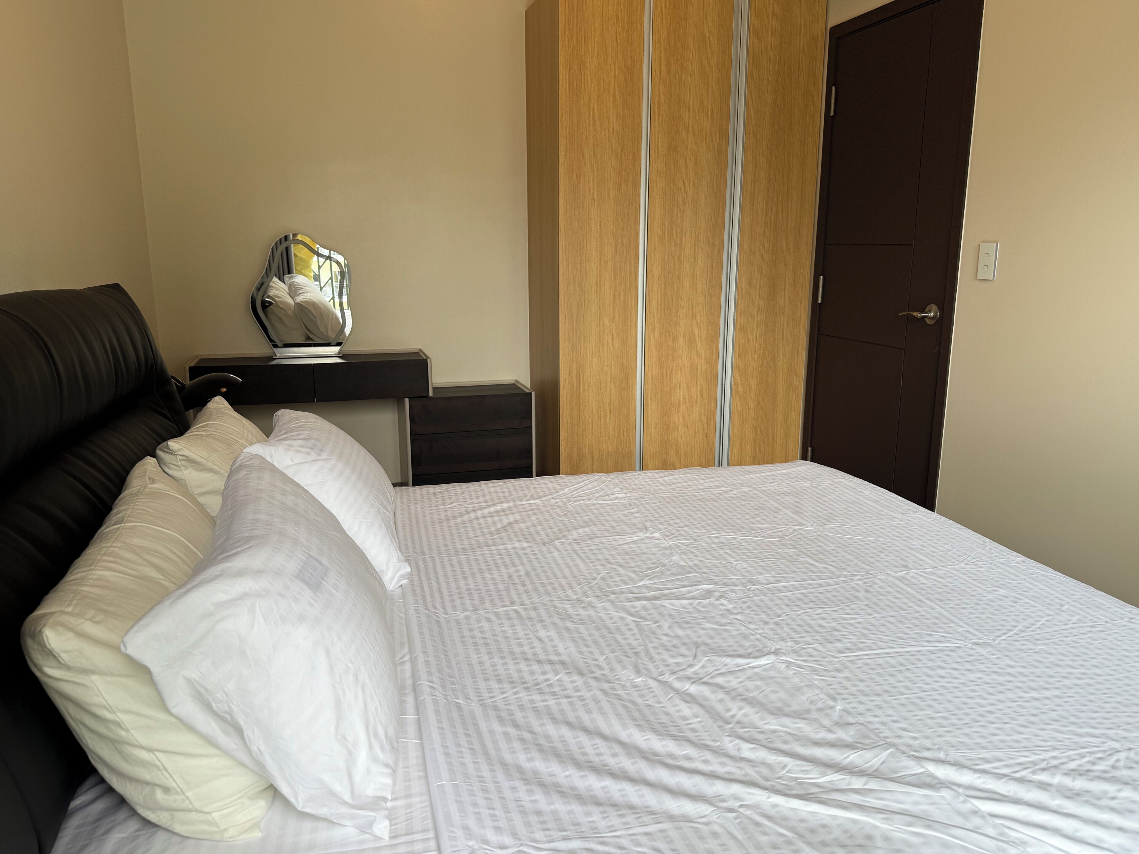 Una cama bien hecha con ropa blanca, un sillón de cuero oscuro, una mesita de noche con una lámpara y un armario de madera.