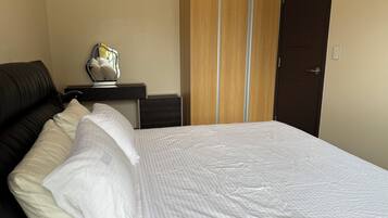 Una cama bien hecha con ropa blanca, un sillón de cuero oscuro, una mesita de noche con una lámpara y un armario de madera.