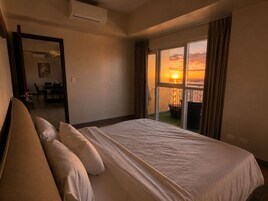 Ein Hotelzimmer mit einem großen Bett, Blick auf den Sonnenuntergang durch das Fenster und einem Balkon mit einem Stuhl.