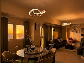 Un salon moderne avec un coin repas, de grandes fenêtres offrant une vue sur le coucher de soleil et des luminaires au design contemporain.