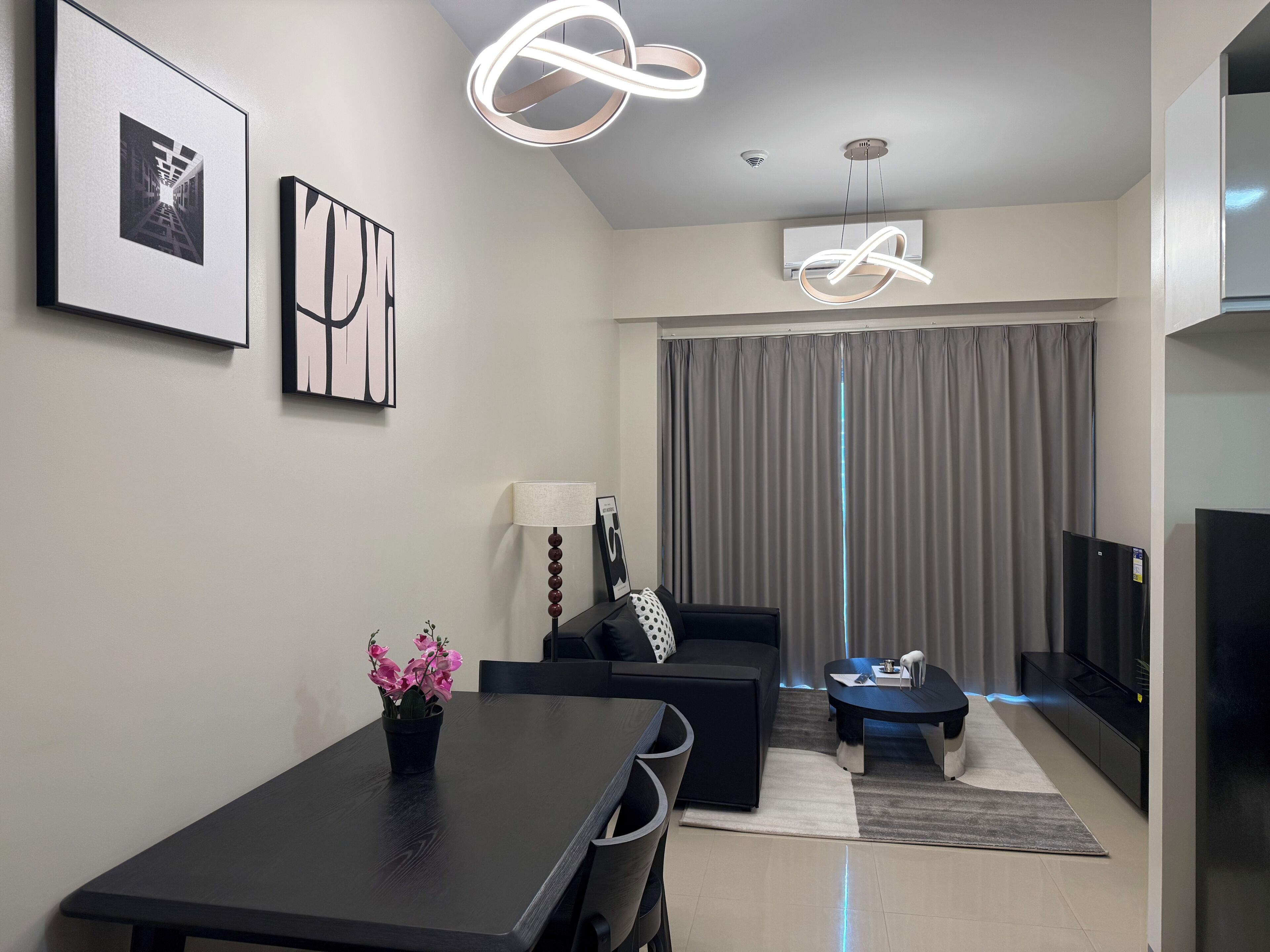 A modern living room with a dining table, sofa, and a TV. There are two framed artworks on the wall, a floor lamp, and a ceiling light fixture.