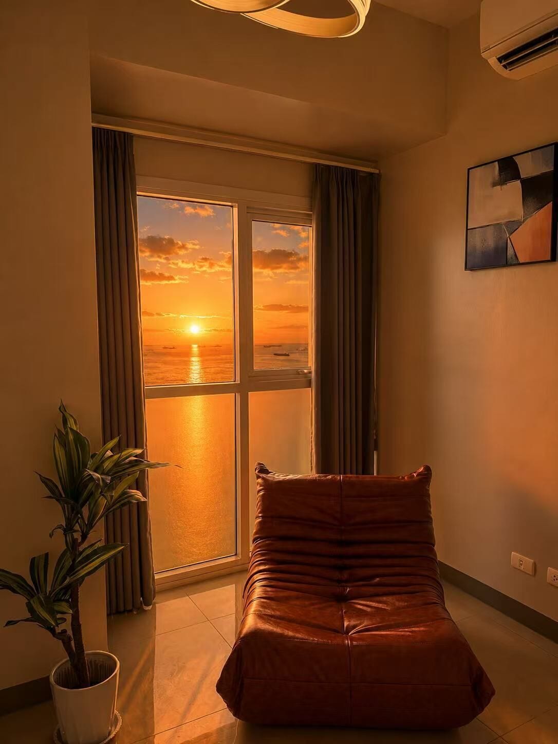 A room with a large window, a brown leather sofa, a potted plant, and a painting on the wall.