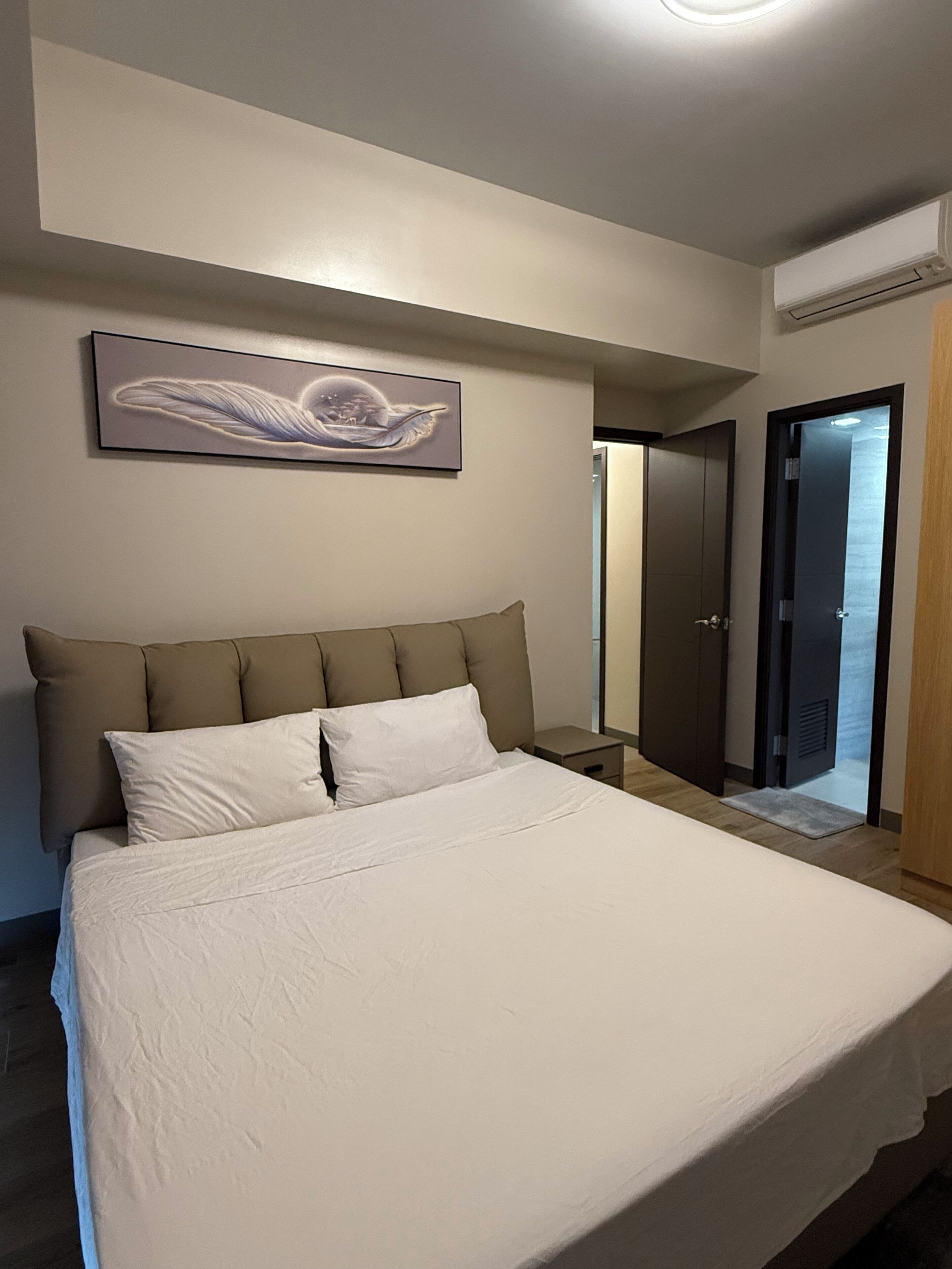 A bedroom with a bed, a nightstand, a wall-mounted artwork, and an air conditioning unit.