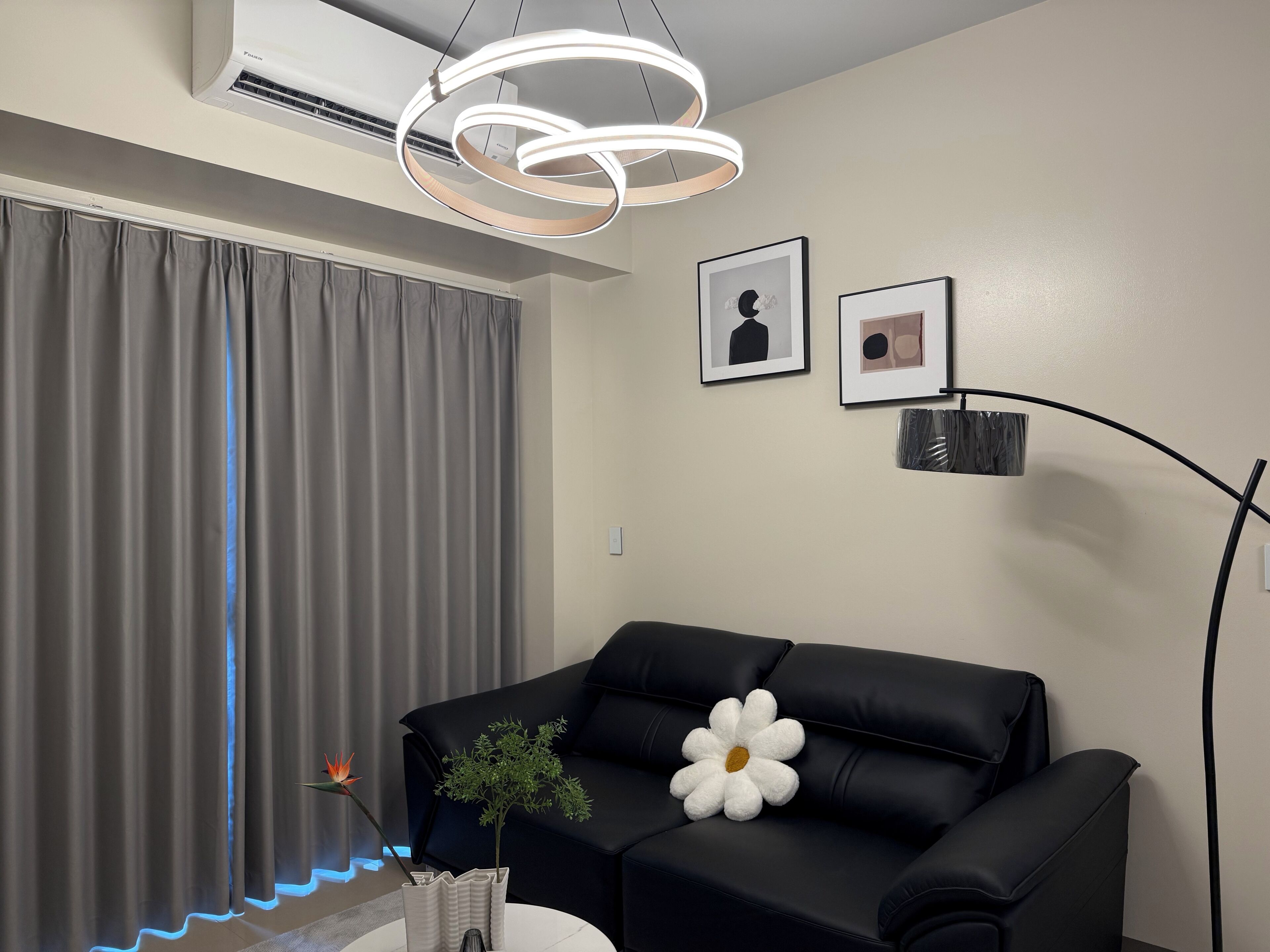 A modern living room with a black leather sofa, a white flower-shaped pillow, a vase with a plant, and a wall-mounted air conditioner.