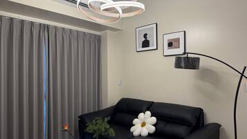 A modern living room with a black leather sofa, a white flower-shaped pillow, a vase with a plant, and a wall-mounted air conditioner.