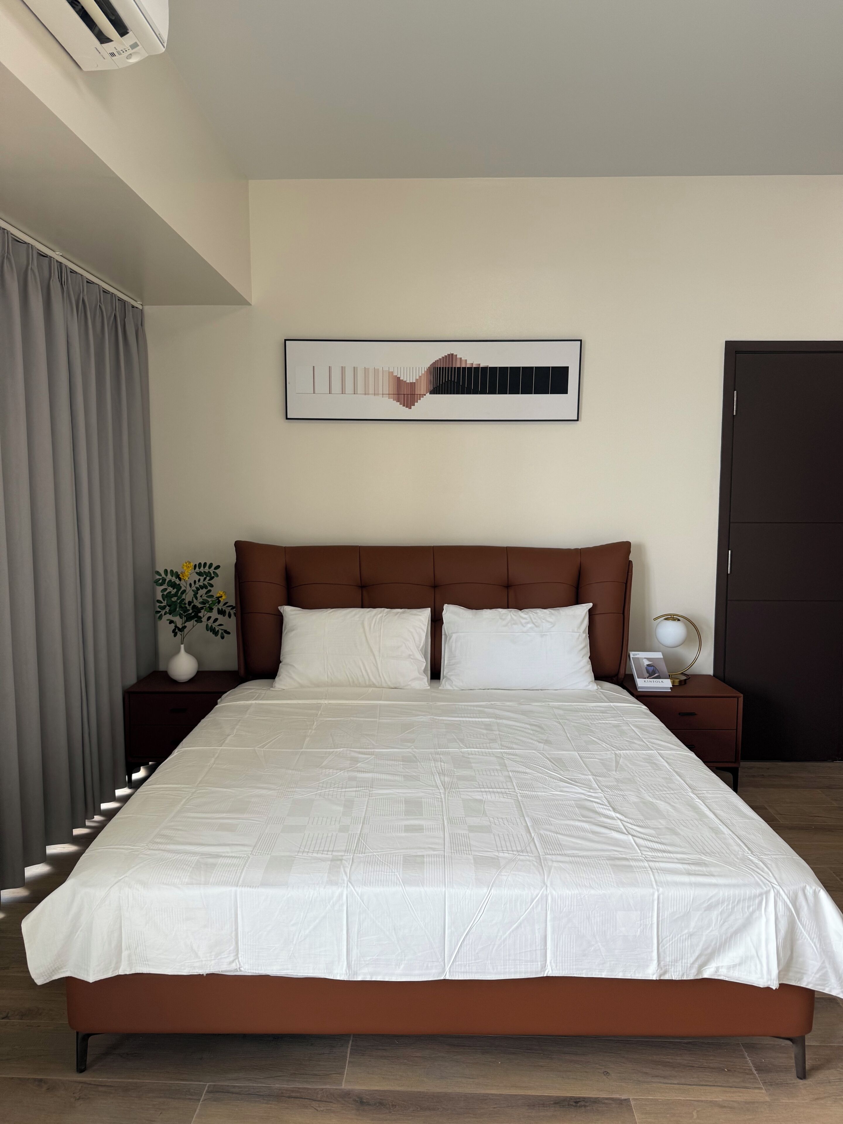 Una camera d'albergo con un letto grande, una testiera, un comodino con una lampada, un quadro incorniciato al muro e una finestra con tende.