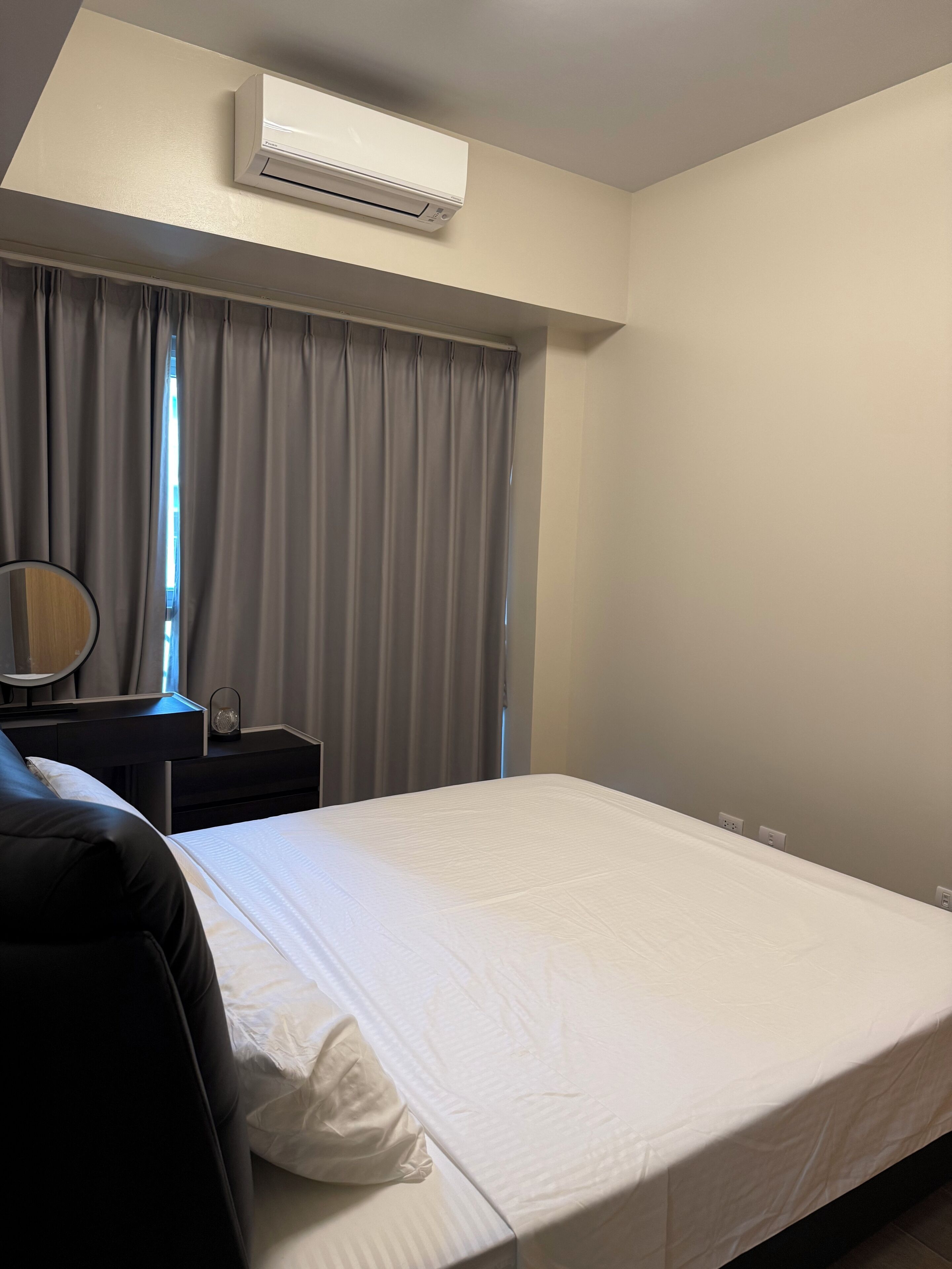A hotel room with a bed, a bedside table with a lamp, a window with curtains, and an air conditioning unit.