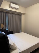 Hotellihuone, jossa on sänky, yöpöytä lampulla, ikkuna verhoilla ja ilmastointilaite.