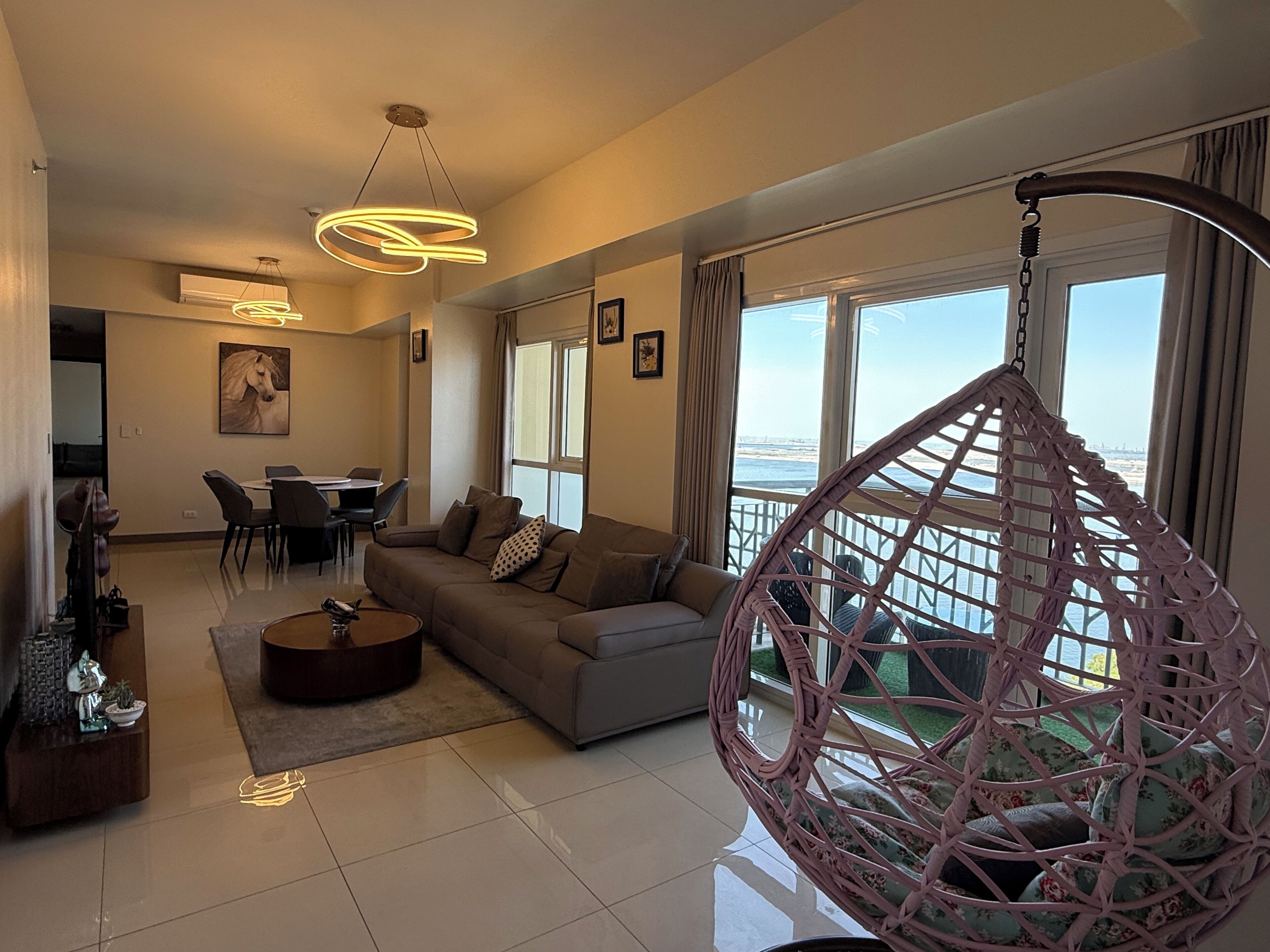 A modern living room with a sofa, coffee table, and hanging chair, a dining area with chairs, and large windows offering a view.