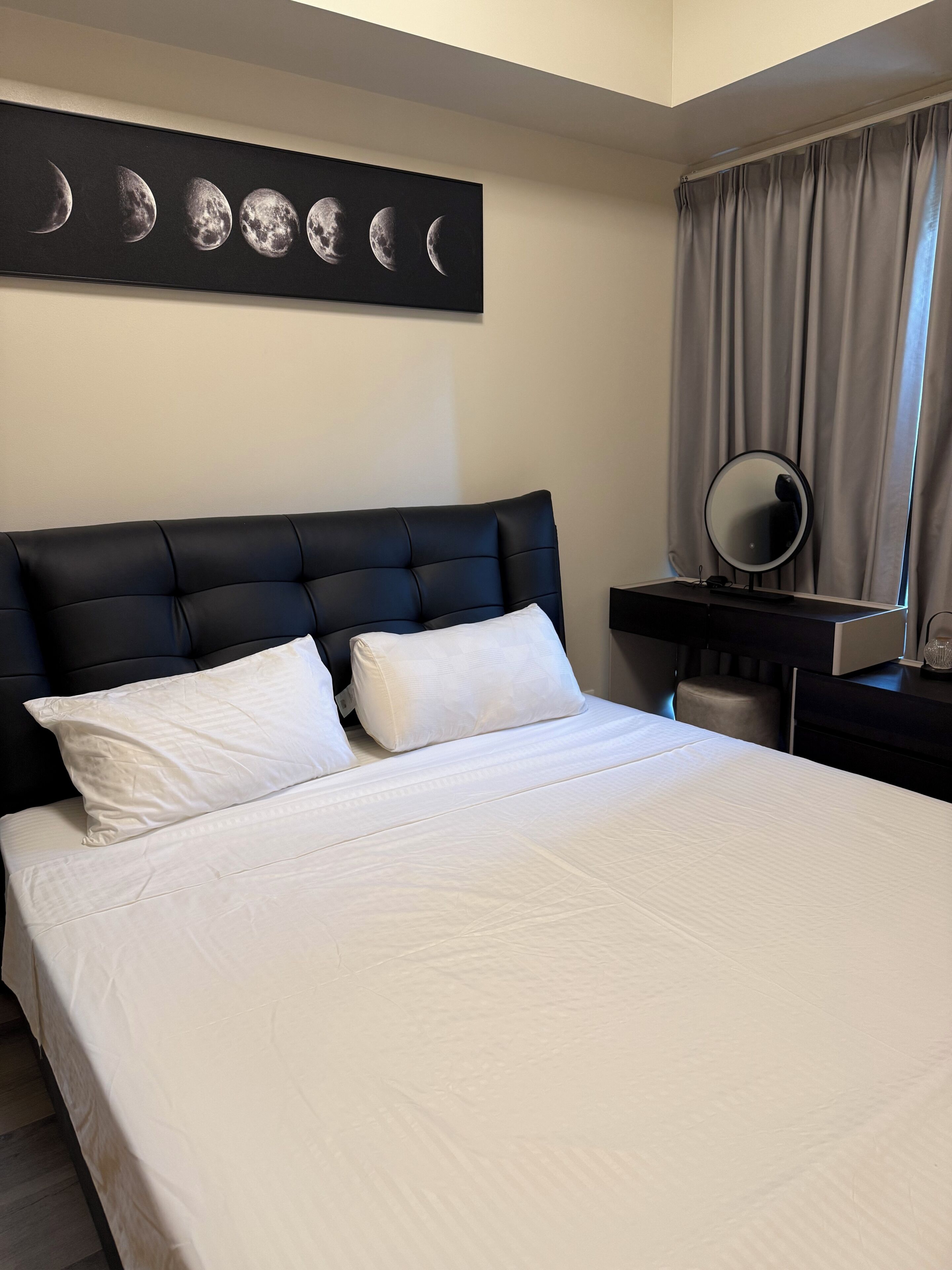 A hotel room with a large bed, a headboard, a wall-mounted artwork of moon phases, a bedside table with a mirror, and a window with curtains.
