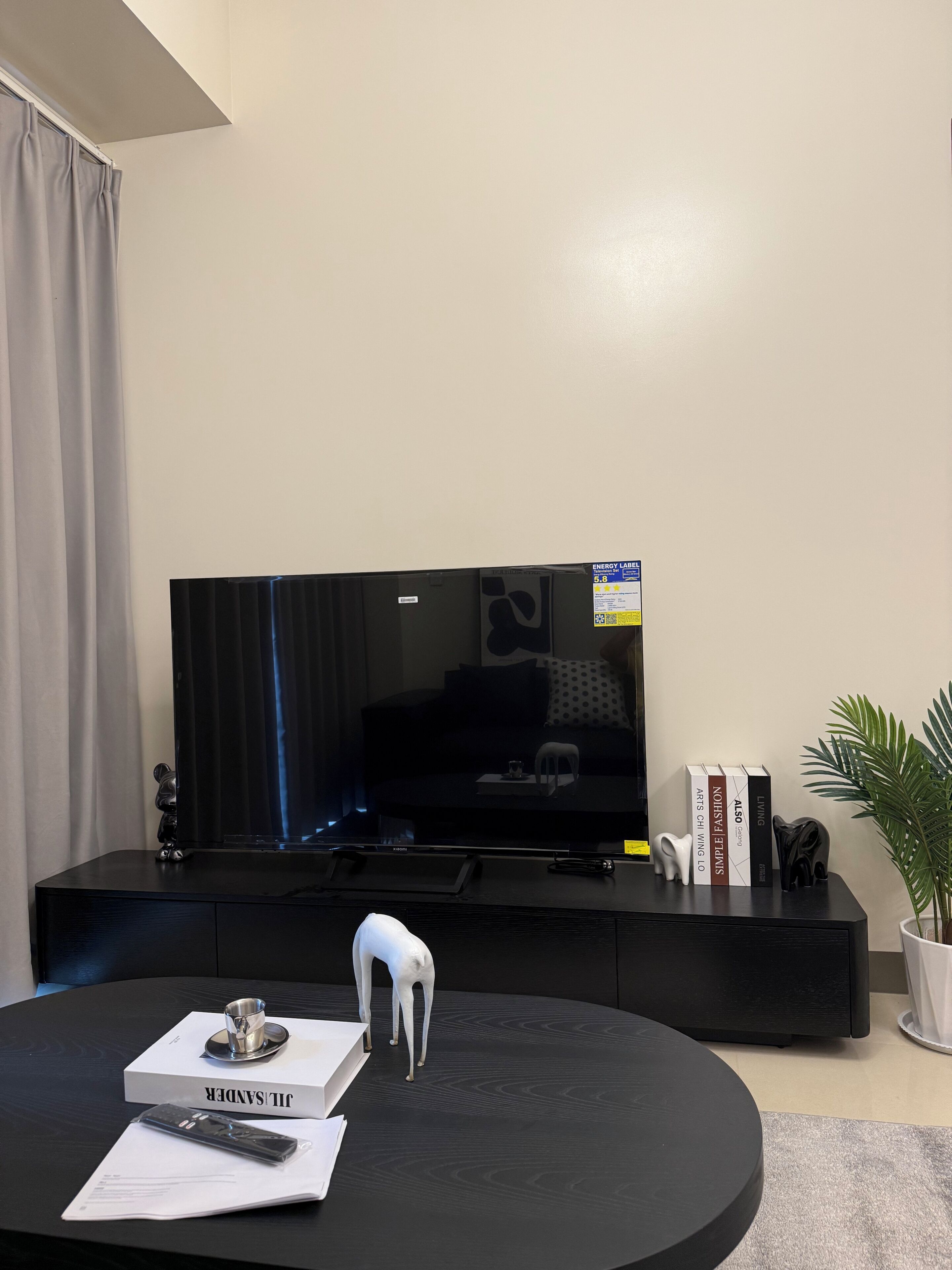 Ett modernt vardagsrum med en platt-TV, en svart möbel för underhållningsutrustning, ett runt soffbord med en fjärrkontroll, en bok och en dekorativ elefantfigur.