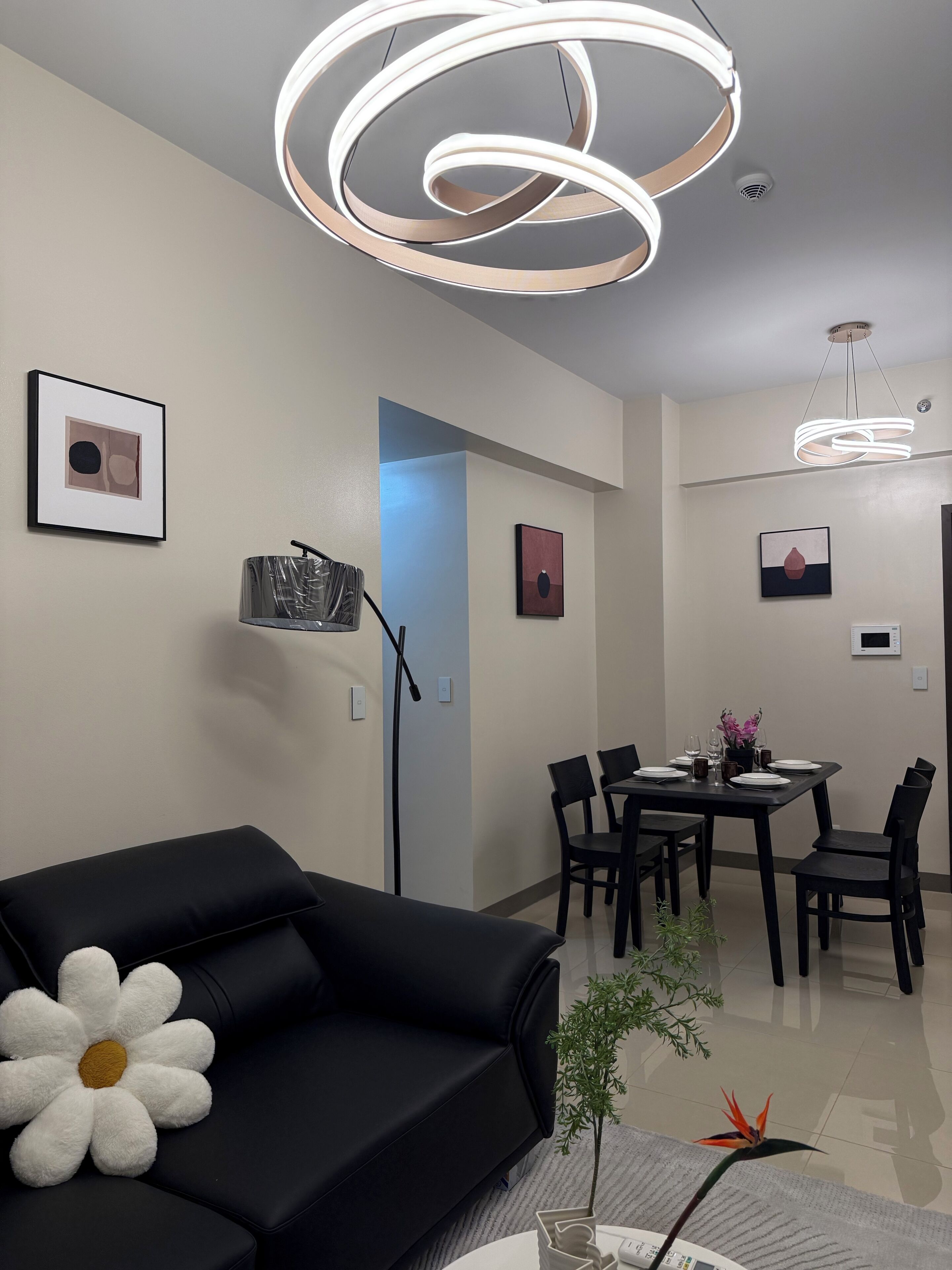 Una sala de estar moderna con un sofá negro, un cojín con forma de flor blanca, una zona de comedor con una mesa preparada para cuatro personas y una lámpara de techo.