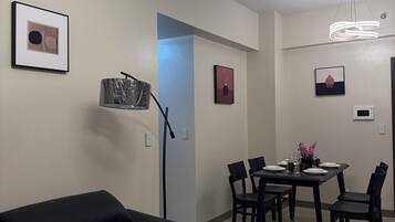 Una sala de estar moderna con un sofá negro, un cojín con forma de flor blanca, una zona de comedor con una mesa preparada para cuatro personas y una lámpara de techo.