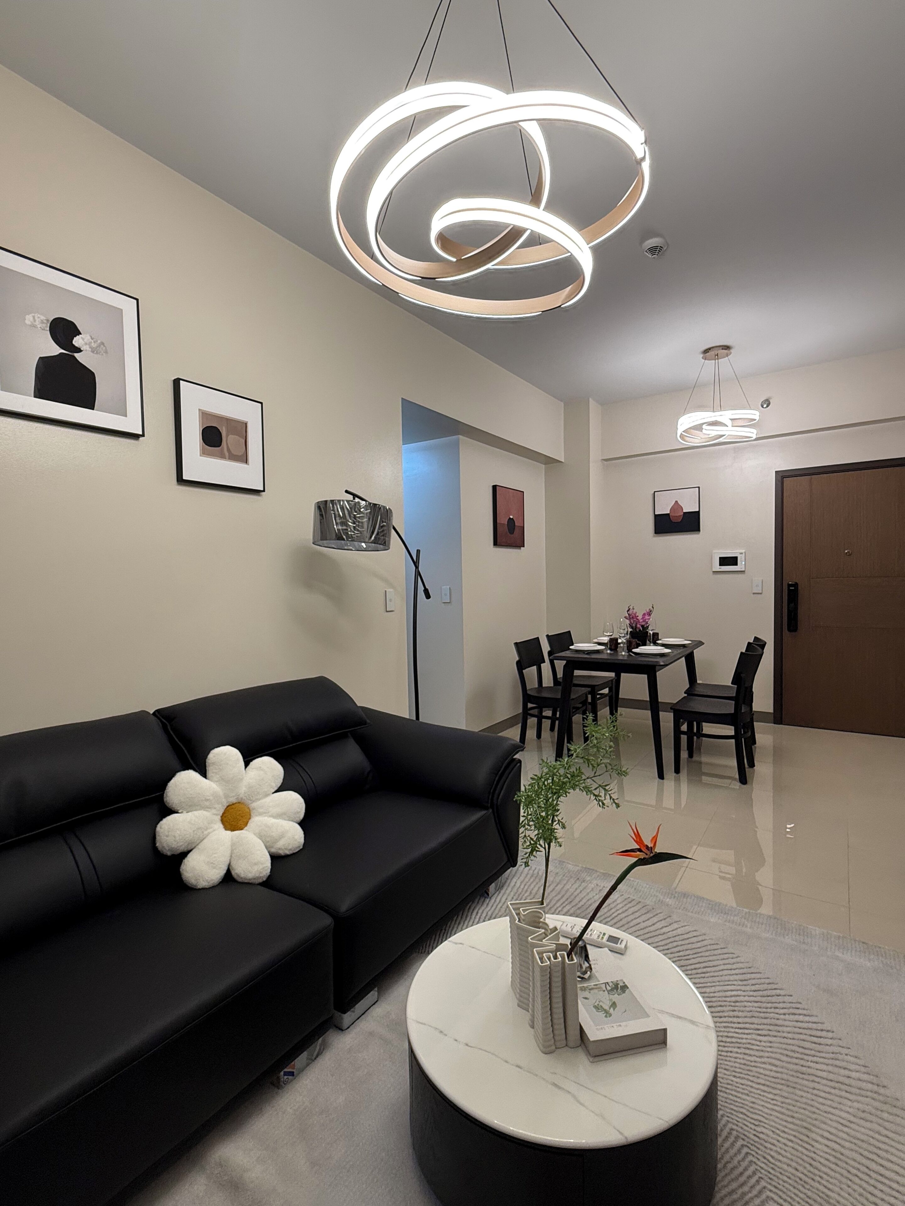 A modern living room with a black sofa, a round coffee table with decorative items, a dining area with a table and chairs, and a hallway leading to a door.