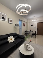 A modern living room with a black sofa, a round coffee table with decorative items, a dining area with a table and chairs, and a hallway leading to a door.