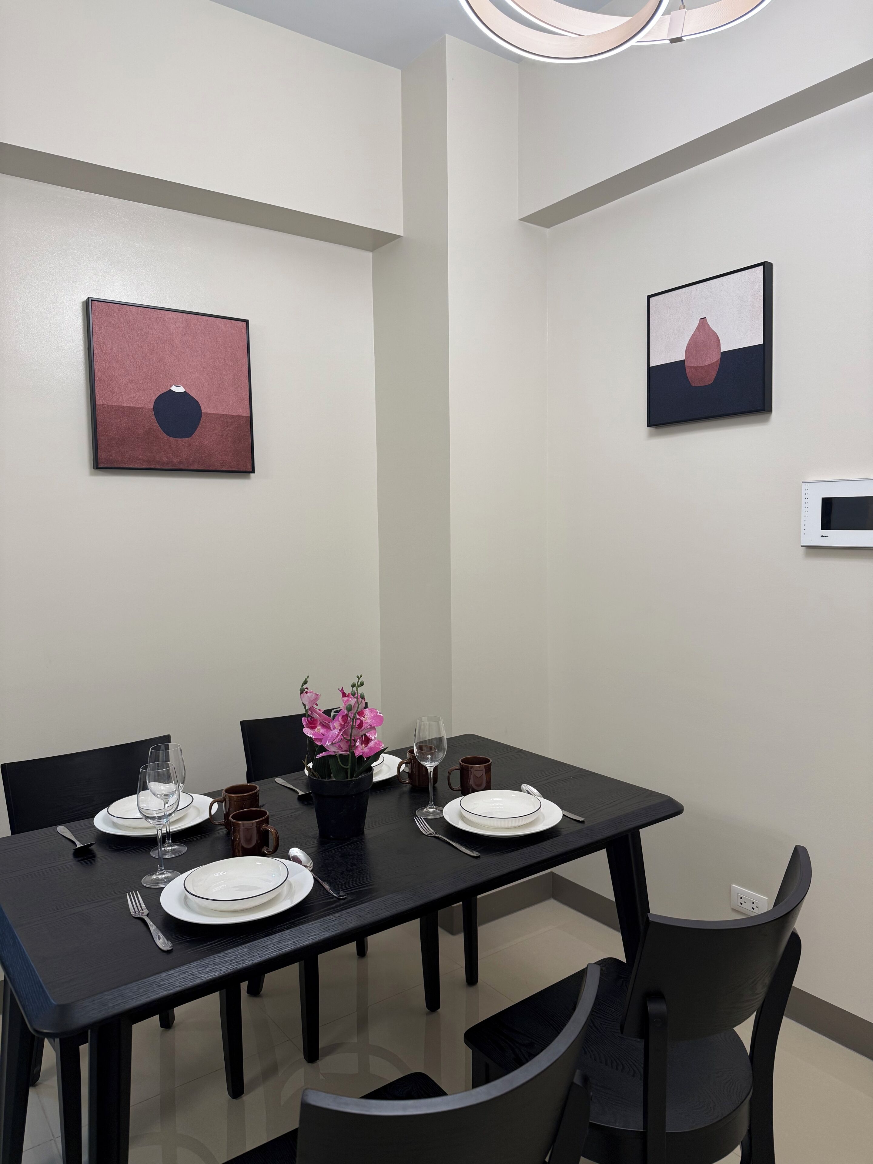 Zona pranzo con tavolo apparecchiato per quattro, comprensivo di piatti, bicchieri e posate. Sono presenti due sedie, una pianta in vaso con fiori rosa e opere d'arte alle pareti.