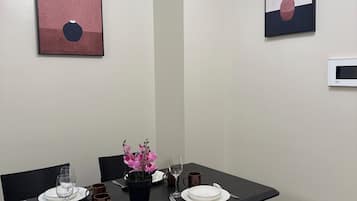 Zona pranzo con tavolo apparecchiato per quattro, comprensivo di piatti, bicchieri e posate. Sono presenti due sedie, una pianta in vaso con fiori rosa e opere d'arte alle pareti.