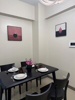 Un comedor con una mesa dispuesta para cuatro, incluyendo platos, vasos y cubiertos. Hay dos sillas, una planta en maceta con flores rosadas y obras de arte en las paredes.