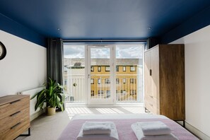 A bedroom with a bed, a wooden dresser, a potted plant, and a view of a building through a glass door. - GuestReady - Gated West London Retreat (NO WIFI) (London)