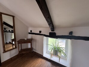 Um quarto com piso de madeira, uma pequena mesa com uma vela, uma janela com uma planta e vigas de madeira aparente no teto.