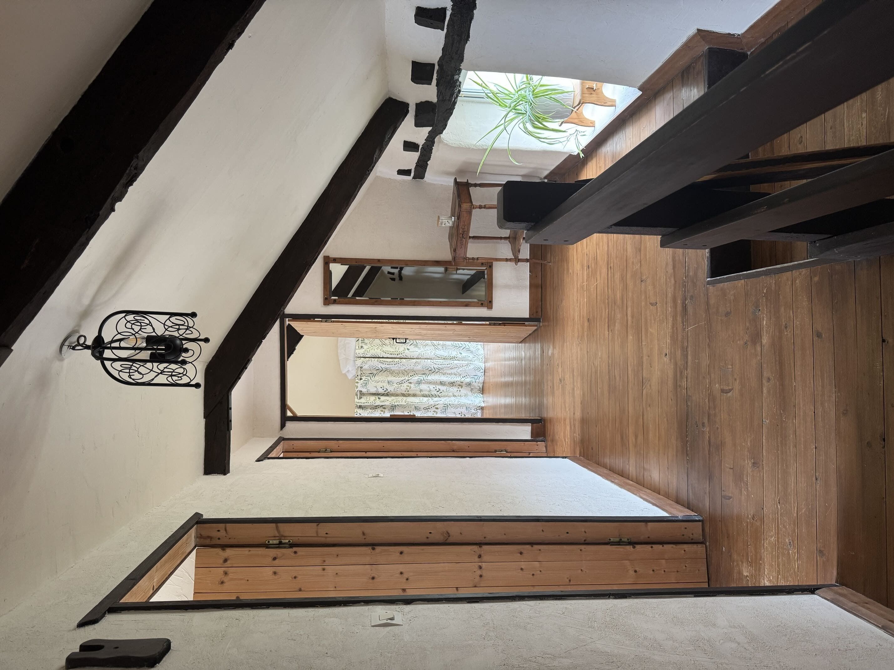 Quarto de sótão rústico, com estrutura de madeira, teto inclinado, pequena janela com planta e luminária fixada na parede.