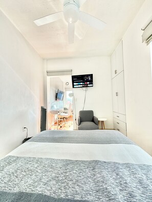 Une chambre à coucher avec un lit, un bureau avec un ordinateur, une chaise, un téléviseur fixé au mur et un ventilateur de plafond.