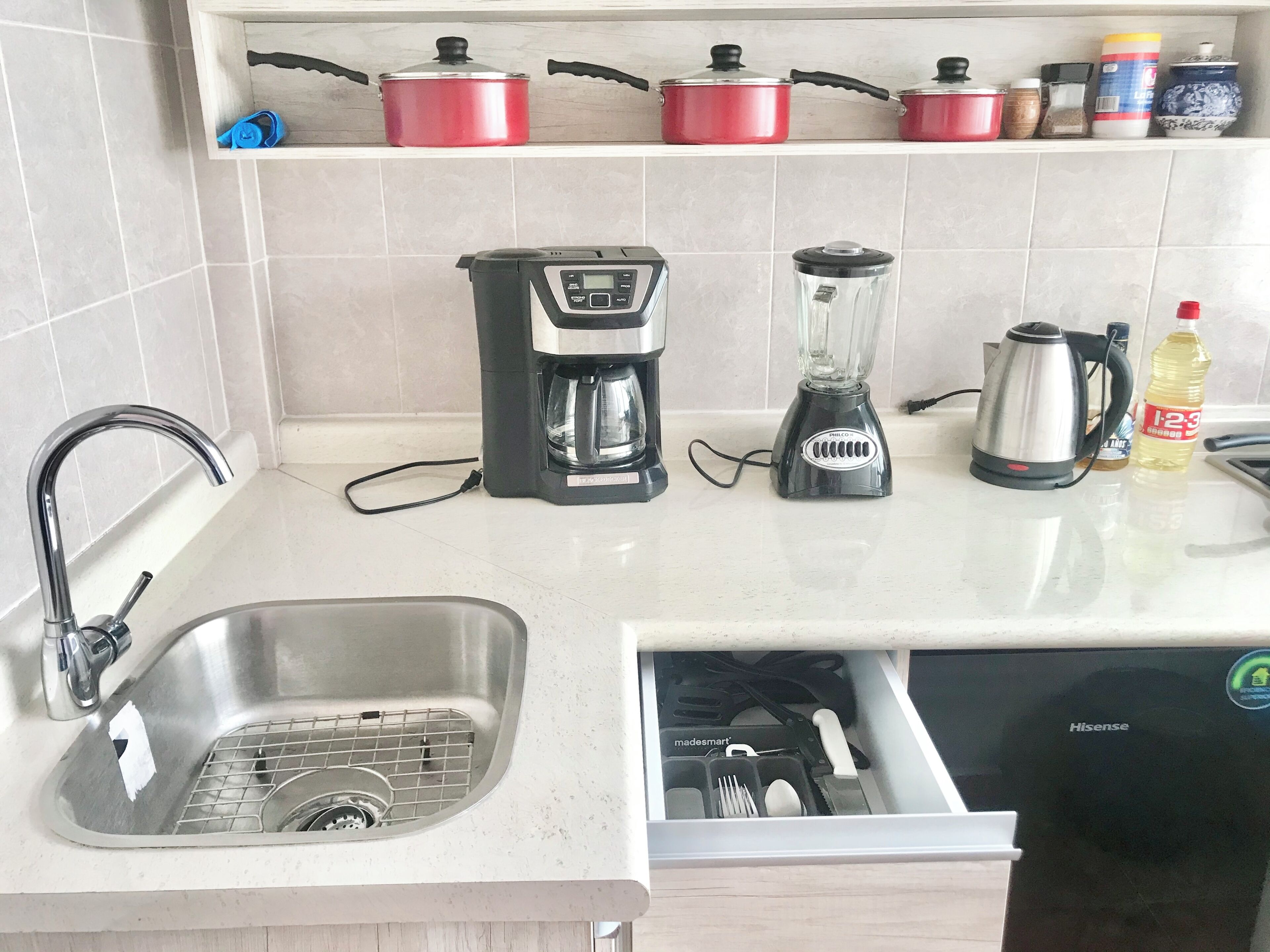 Una cocina con un lavamanos, una cafetera, una licuadora, un hervidor y un cajón con utensilios.