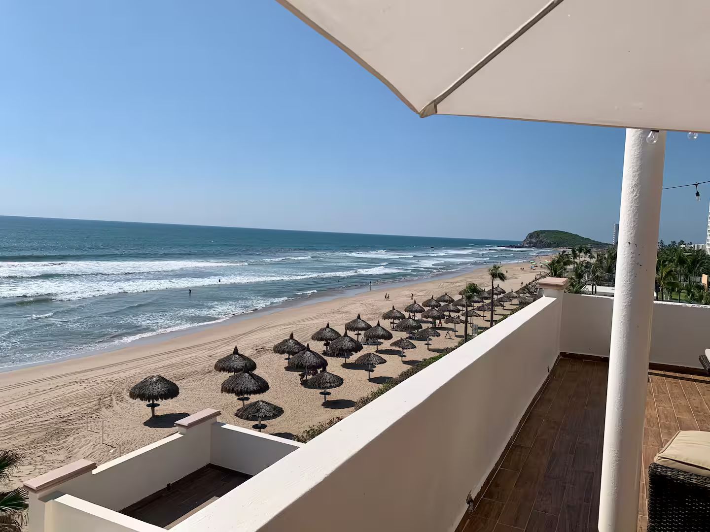 Een balkon met uitzicht op een strand, rieten parasols en een heldere lucht.