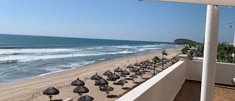 A balcony with a view of a beach, thatched umbrellas, and a clear sky.